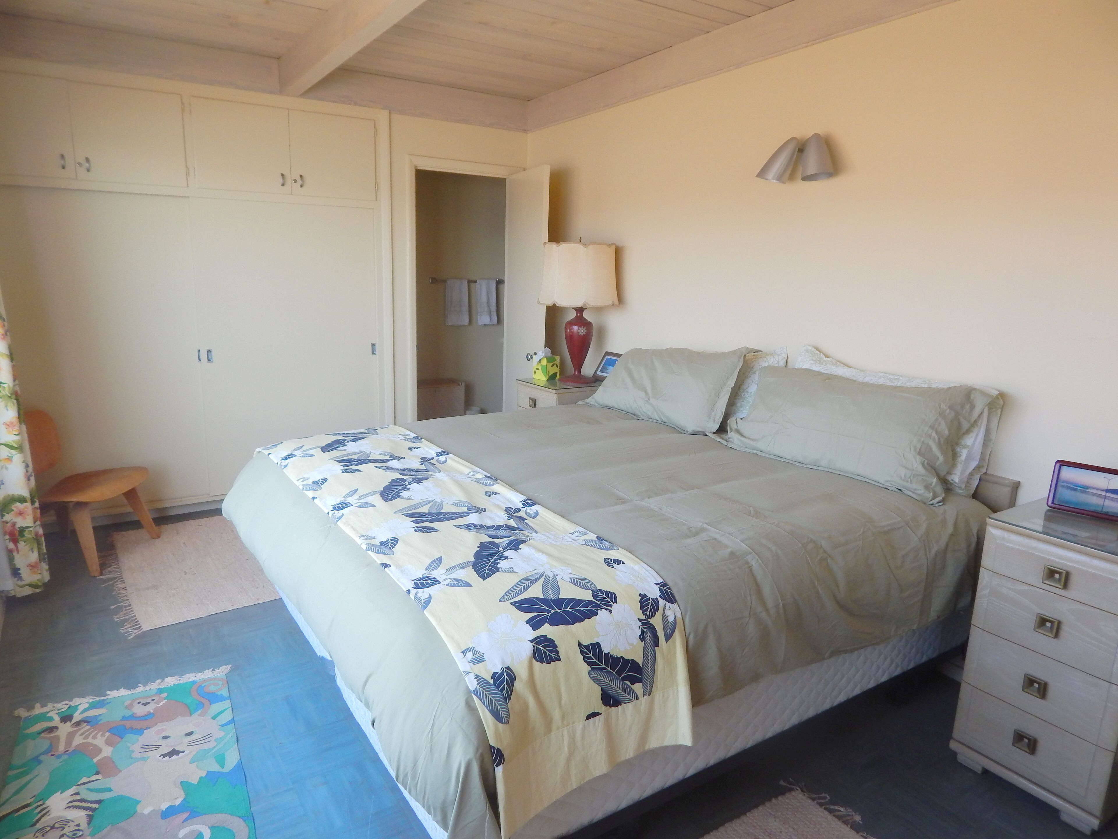 Quarto com cama, mesa de cabeceira, luminária, cadeira, guarda-roupa e banheiro visível por uma porta aberta.