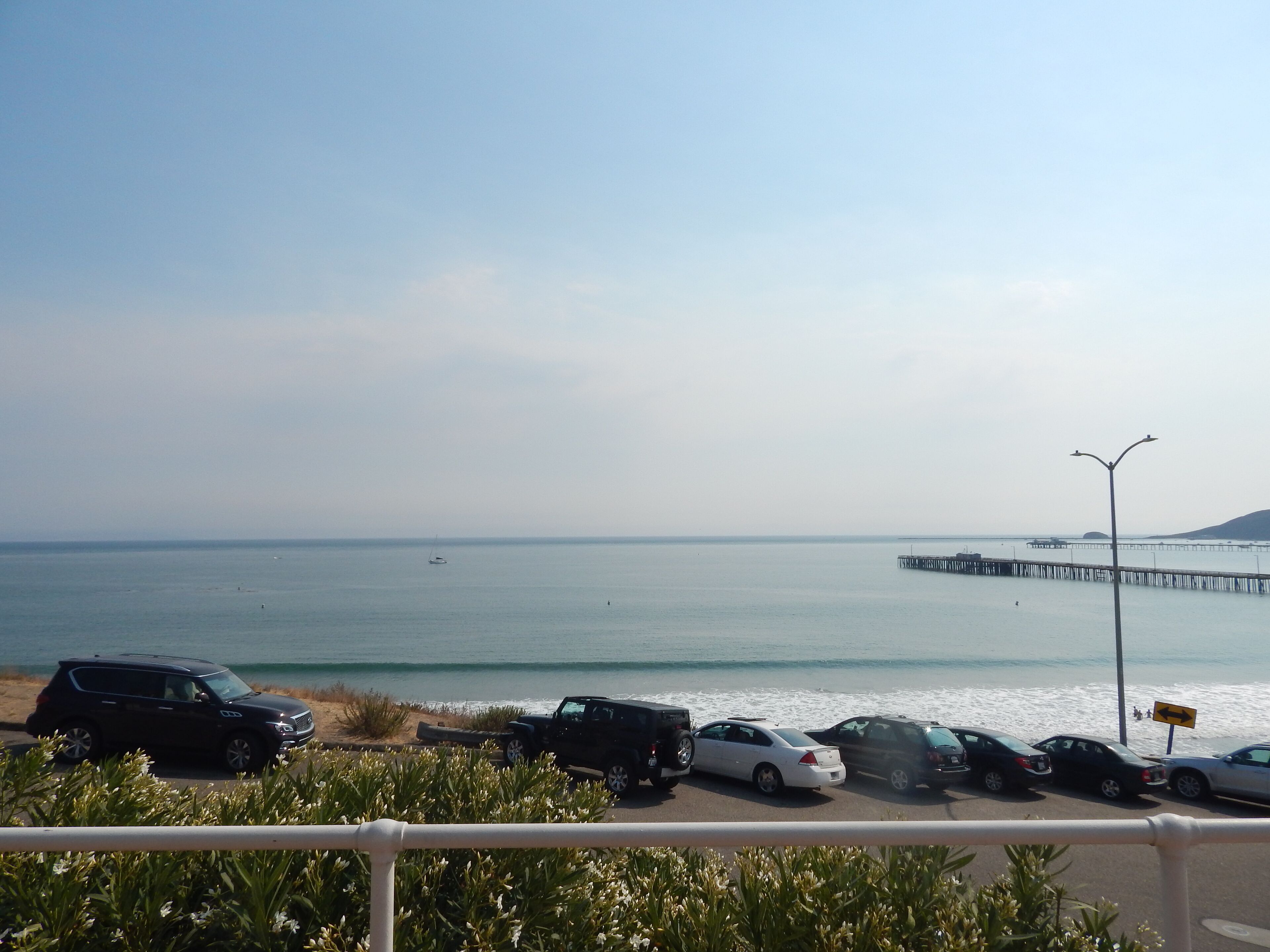 Ein Küstenparkplatz mit Autos, einer Pier, die sich ins Wasser erstreckt, und einem wolkenlosen Himmel.