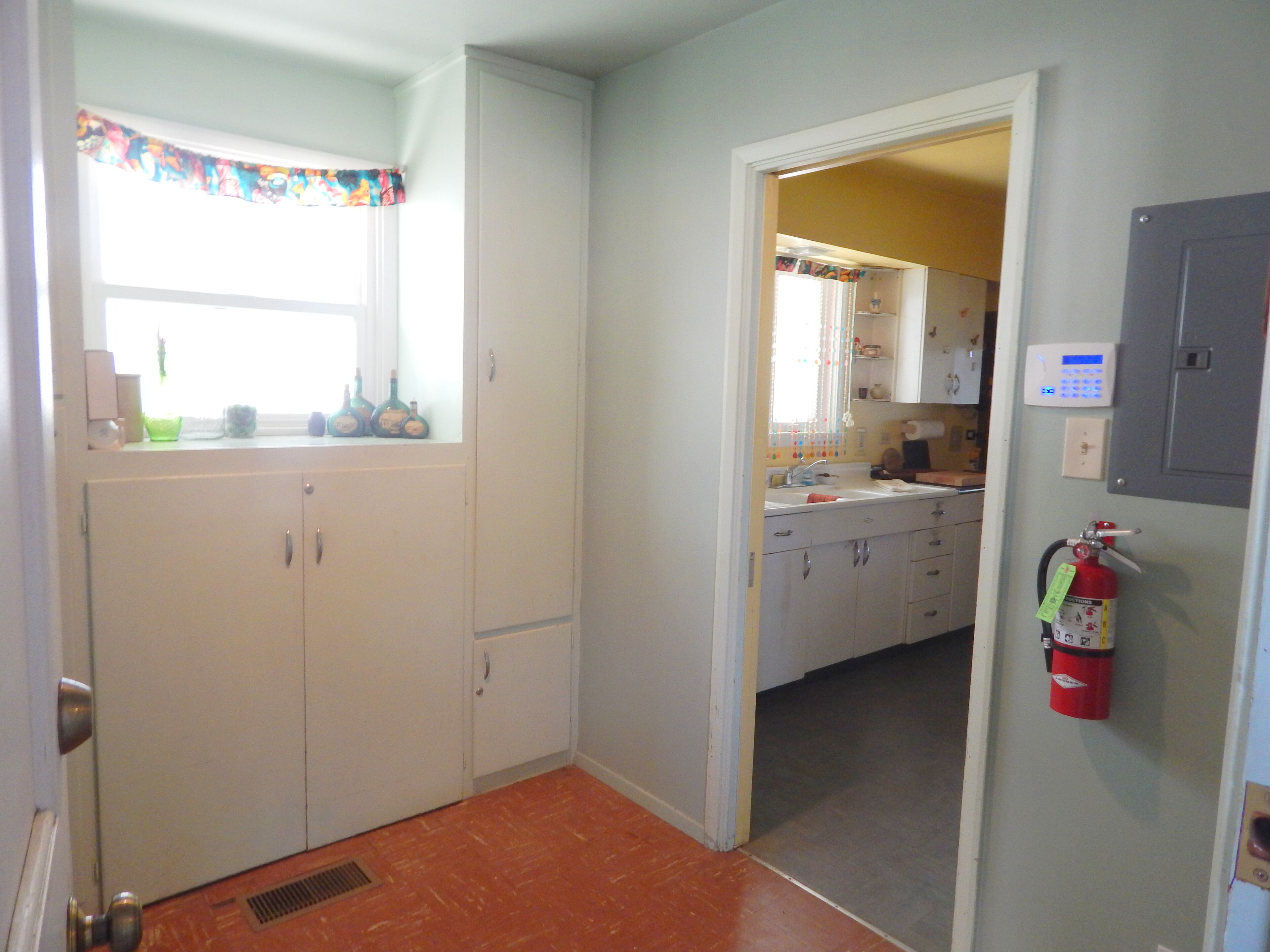 A kitchen with white cabinets, a window with colorful curtains, a fire extinguisher mounted on the wall, and a door leading to another room.
