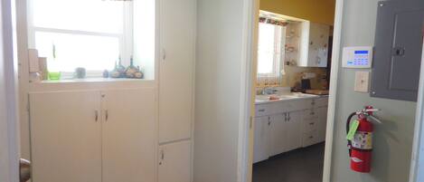 A kitchen with white cabinets, a window with colorful curtains, a fire extinguisher mounted on the wall, and a door leading to another room.