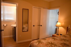 A bedroom with a bed, a nightstand, a lamp, a window with blinds, a door, and a bathroom with a sink and toilet.