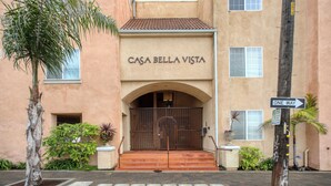 The entrance to a building named "Casa Bella Vista" with a metal gate, a "One Way" sign, and a palm tree in the foreground. - 210 Casa Bella: 2  BR, 2  BA Condominium in Pismo Beach, Sleeps 6 (Pismo Beach)
