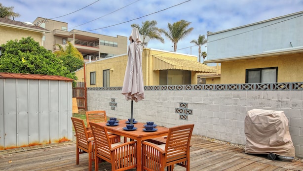 A wooden deck with a table and chairs, a covered grill, and a white wall with decorative tiles. - 369 Stimson: 3 BR, 2 BA House in Pismo Beach, Sleeps 8 (Pismo Beach)
