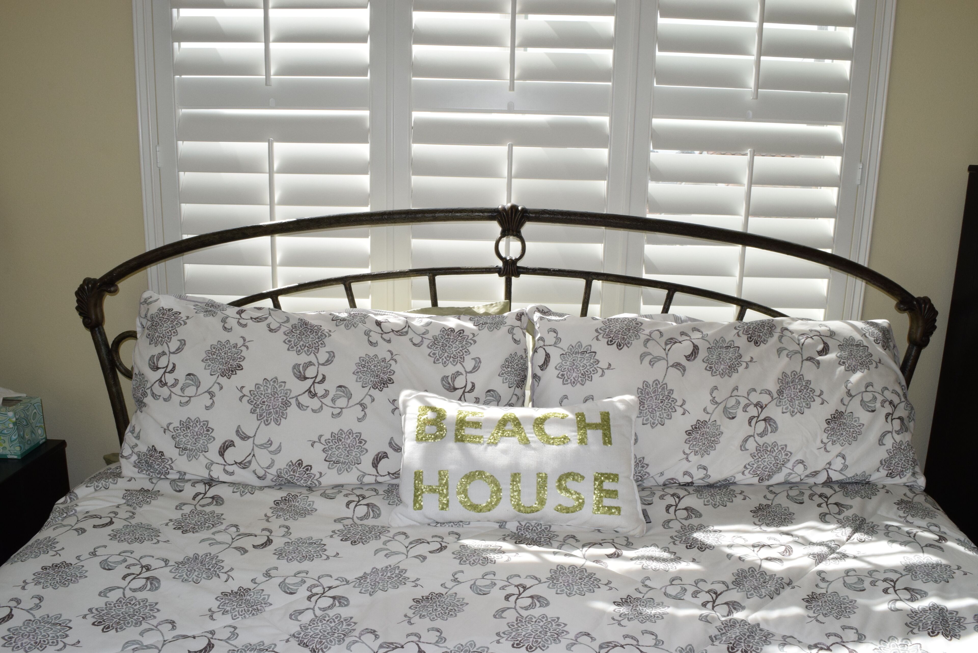 A bed with a metal frame, white bedding with a floral pattern, and a pillow with the text "BEACH HOUSE".