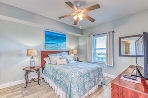 Ein Schlafzimmer mit einem Bett, einem Deckenventilator, einem Nachttisch, einer Lampe, einem Fernseher und einem Fenster mit Vorhängen.