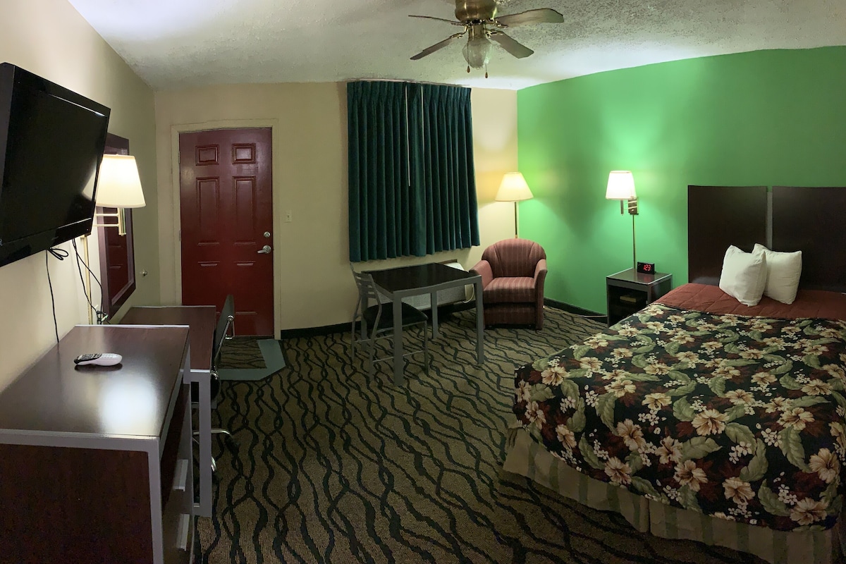 Habitación de hotel con cama, escritorio, silla, televisor y ventilador de techo.