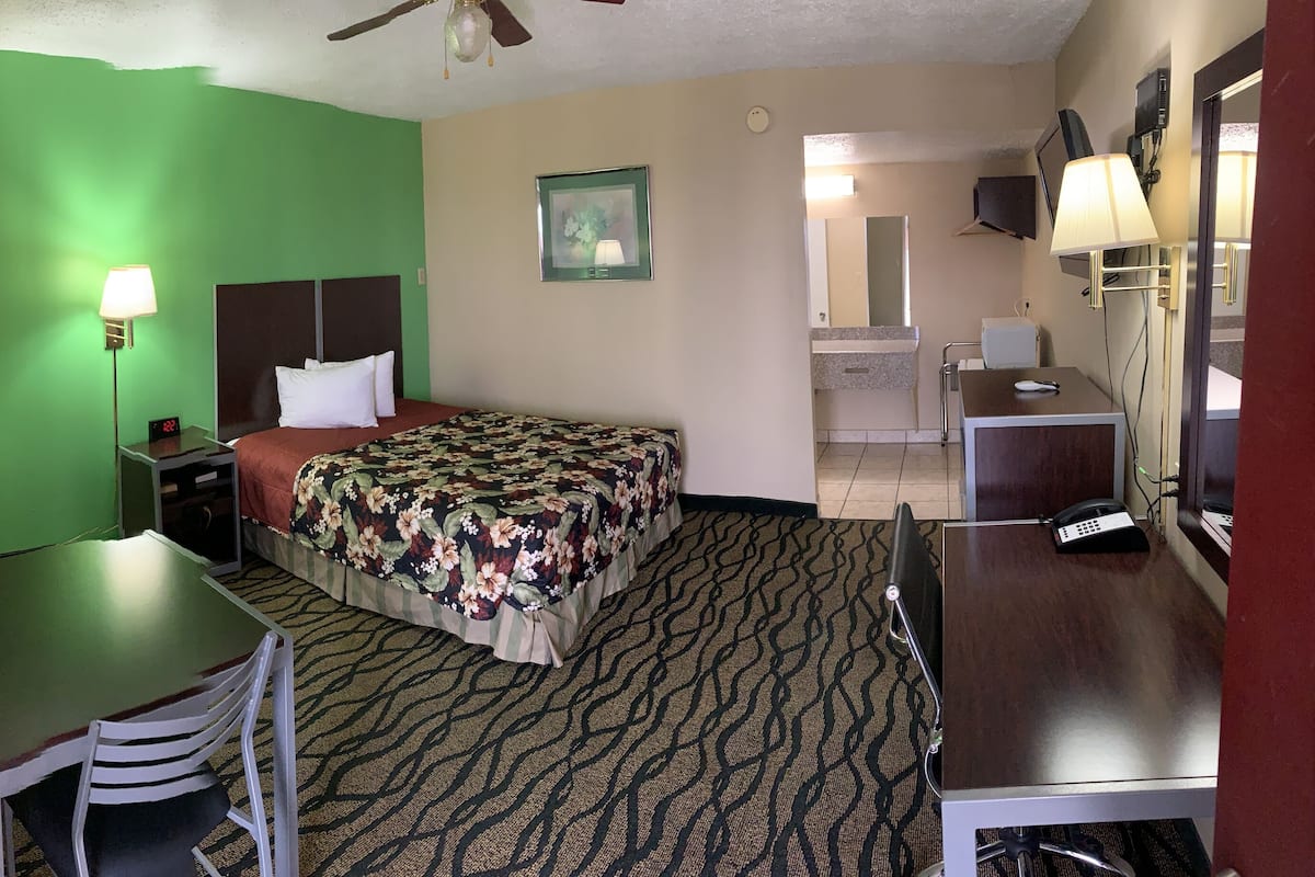 A hotel room with a green wall, a bed with a floral bedspread, a desk with a chair, a small table, a nightstand with a lamp, a TV, and a telephone.
