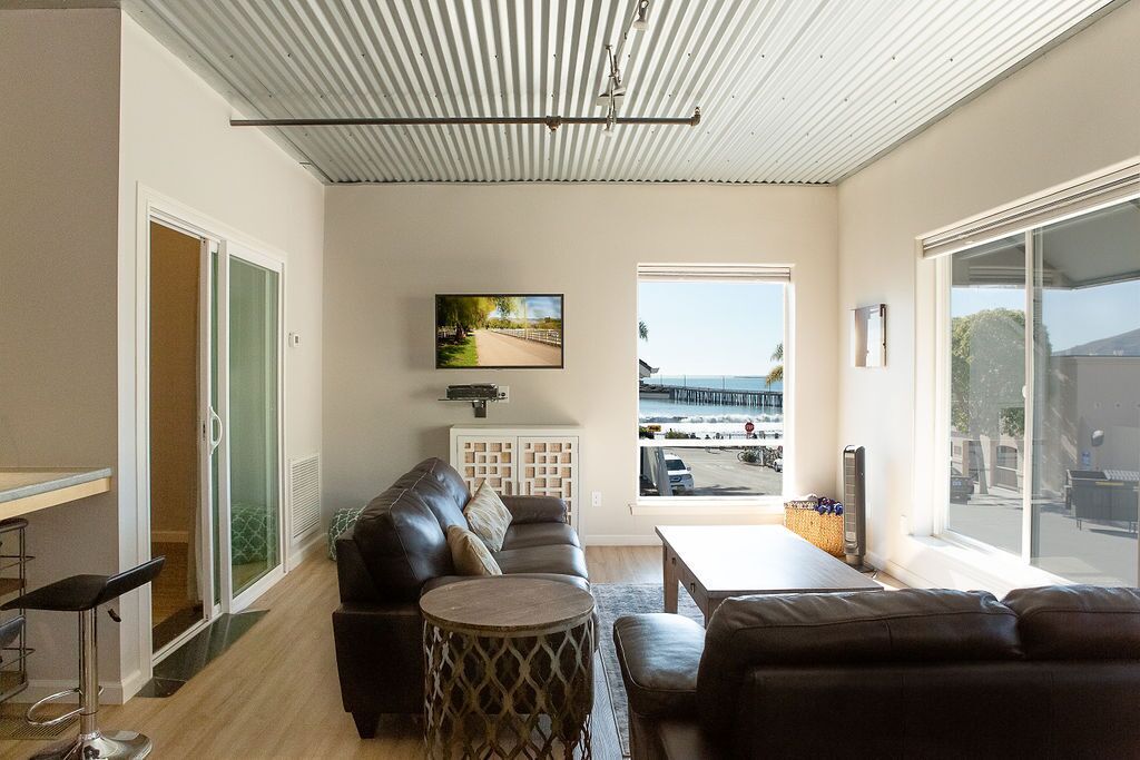 Een moderne woonkamer met een leren bank, een ronde salontafel en een aan de muur bevestigde televisie. Groot raam met uitzicht op het strand en de pier.