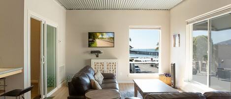 Een moderne woonkamer met een leren bank, een ronde salontafel en een aan de muur bevestigde televisie. Groot raam met uitzicht op het strand en de pier.