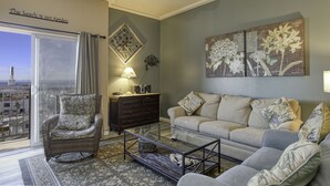 Un salon avec un fauteuil en osier, un canapé, une table basse en verre et une vue sur la ville, visible à travers une grande fenêtre.