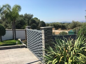 A gated outdoor area with a view of distant mountains, a concrete pathway, and a variety of trees.