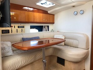 Interior compacto de uma motorhome, com mesa de madeira, assentos acolchoados, painel de controle e uma pequena janela com cortinas.