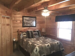 Une chambre dans une cabane en bois, avec un lit, un ventilateur de plafond, un tableau encadré au mur et une fenêtre avec des stores.