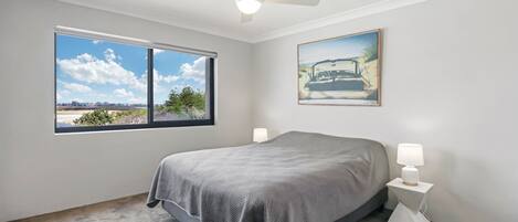 A bedroom with a bed, a ceiling fan, a window with a city view, and a painting on the wall.