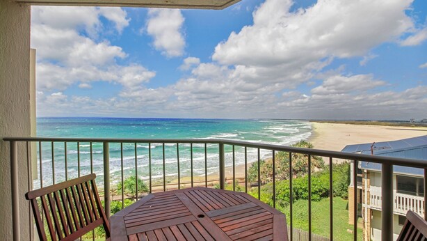 Un balcon doté d’une table et de chaises en bois, donnant sur une plage aux eaux turquoise et au rivage sablonneux.