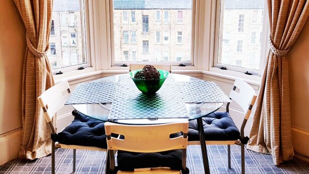 Ein Eckzimmer mit einem runden Glastisch, zwei Stühlen und Blick auf Gebäude durch große Fenster.