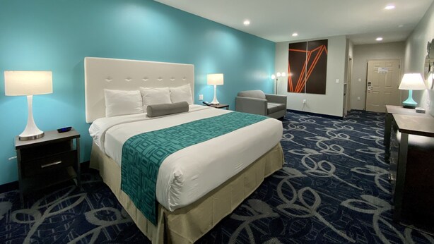 Una habitación de hotel con una cama grande, dos mesitas de noche con lámparas, una silla, un escritorio con lámpara y una puerta.