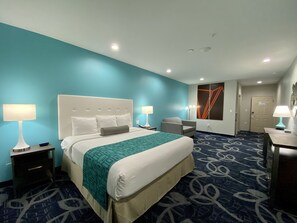 Ein Hotelzimmer mit einem großen Bett, zwei Nachttischen mit Lampen, einem Sessel, einem Schreibtisch mit Lampe und einer Tür.