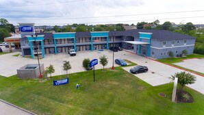 Un edificio de varios pisos con una combinación de colores azul y gris, que cuenta con un estacionamiento con varios autos, un área ajardinada con árboles pequeños y un letrero que indica el nombre 'AMERICAS'.