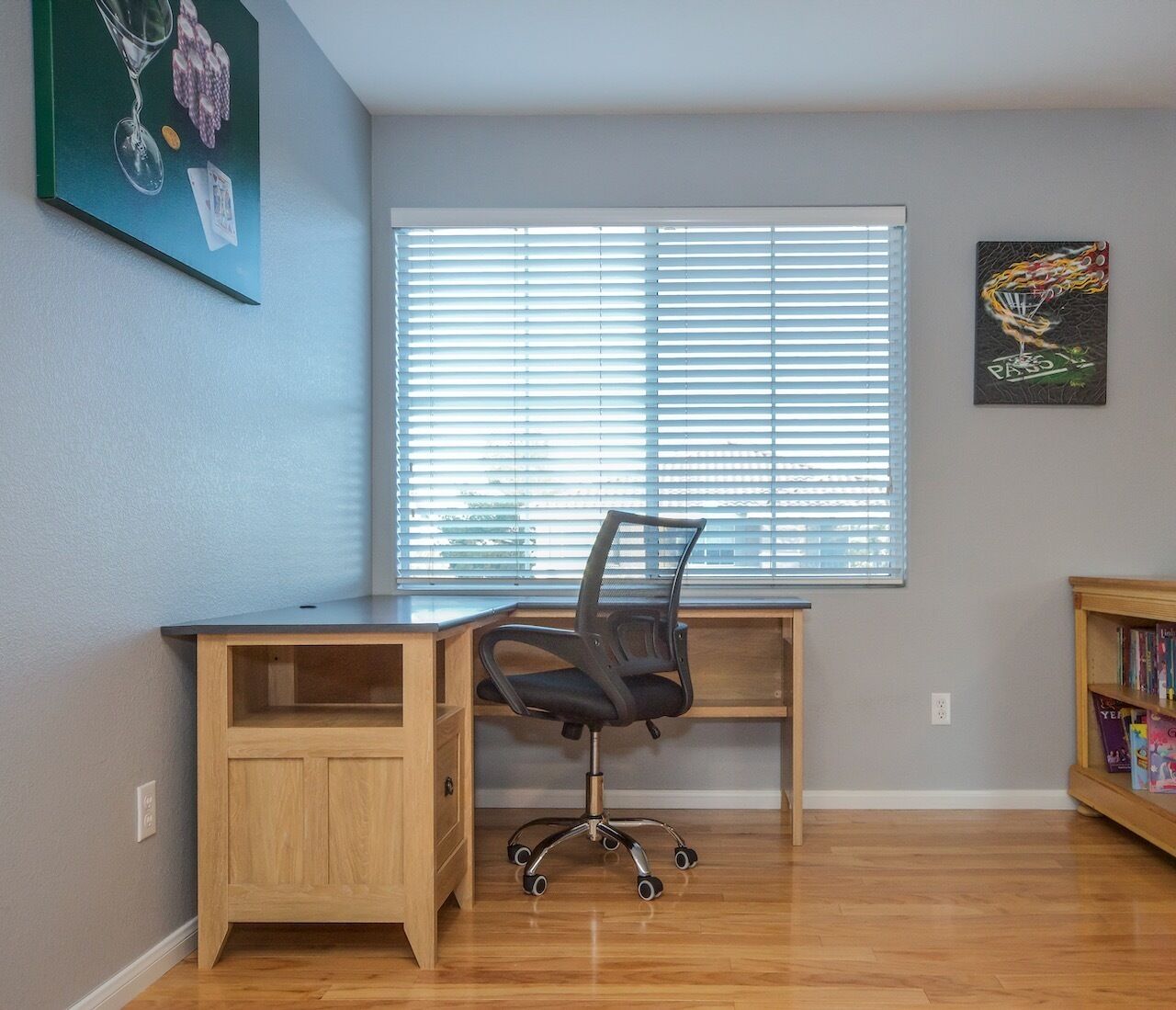 A room with a wooden desk, a black office chair, a window with blinds, and a bookshelf.