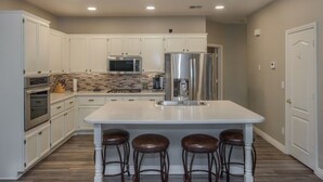 Une cuisine moderne avec des meubles blancs, un grand îlot central doté de tabourets, des appareils en acier inoxydable et un pare-feu en carreaux.