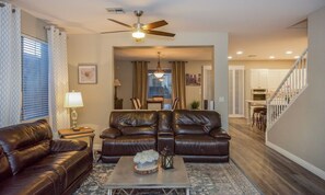 A living room with a brown leather sofa, a coffee table with decorative items, a ceiling fan, and a view into the kitchen.