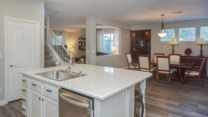 Une cuisine dotée d’un plan de travail blanc, d’un évier en acier inoxydable et d’un lave-vaisselle encastré. On y trouve également un coin repas avec des chaises et une table en bois.