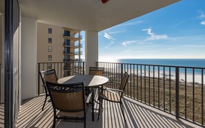A balcony with a table and chairs, overlooking a beach and ocean.