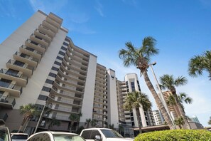 A high-rise building with multiple balconies, palm trees, and parked cars.