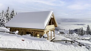 A wooden cabin with a snow-covered roof, surrounded by snow-covered trees and a mountainous landscape.