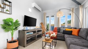 A modern living room with a grey sofa, a flat-screen TV, a wooden cabinet, and a large window with curtains.