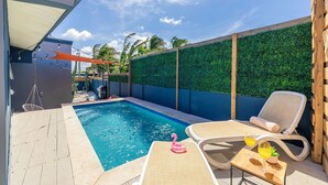 A swimming pool with lounge chairs and a table, surrounded by a wooden deck and a green hedge wall.