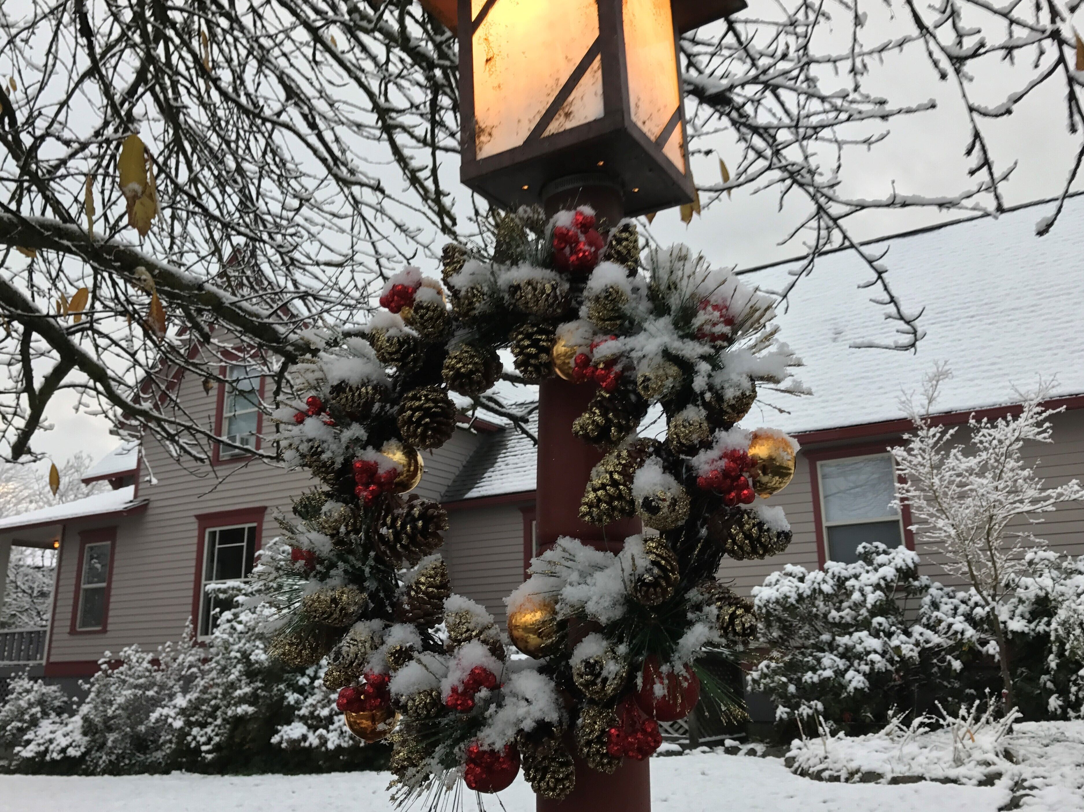 A wreath decorated with red and gold ornaments, pine cones, and greenery, hanging on a post in front of a house with a snow-covered roof.