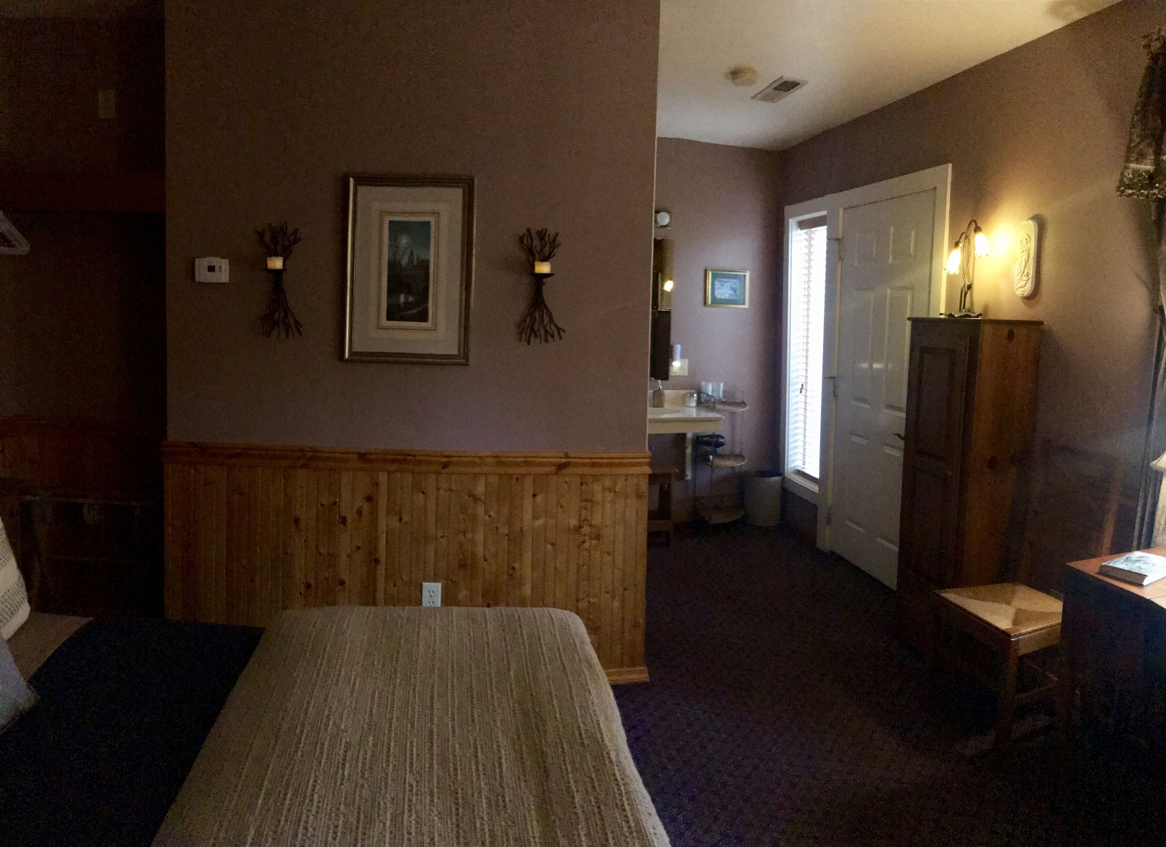 A hotel room with a bed, wooden paneling, a small desk, a mirror, and a window with blinds.