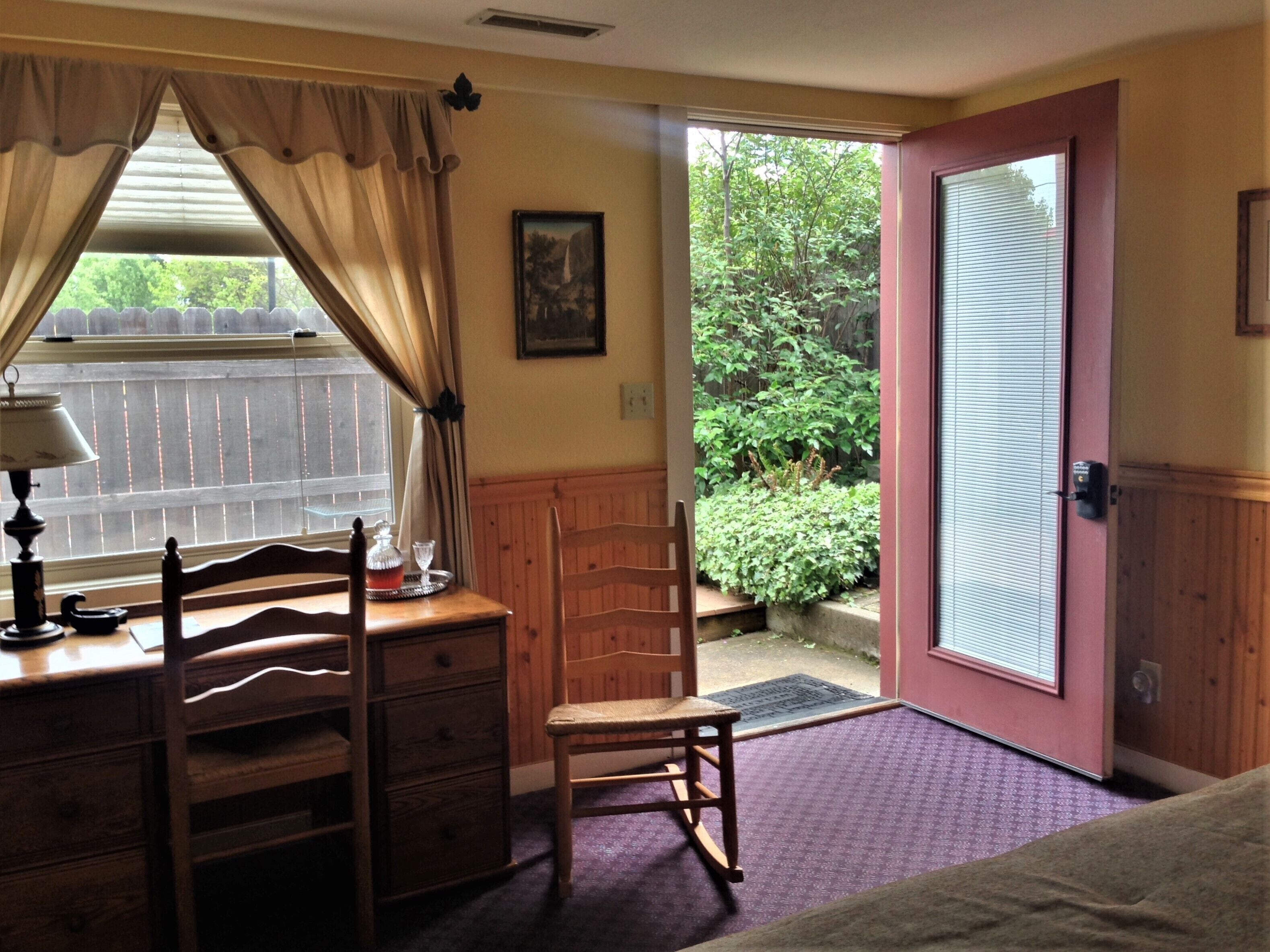 A room with a wooden desk, chair, and a bed. There is a window with curtains, a door leading outside, and a framed picture on the wall.