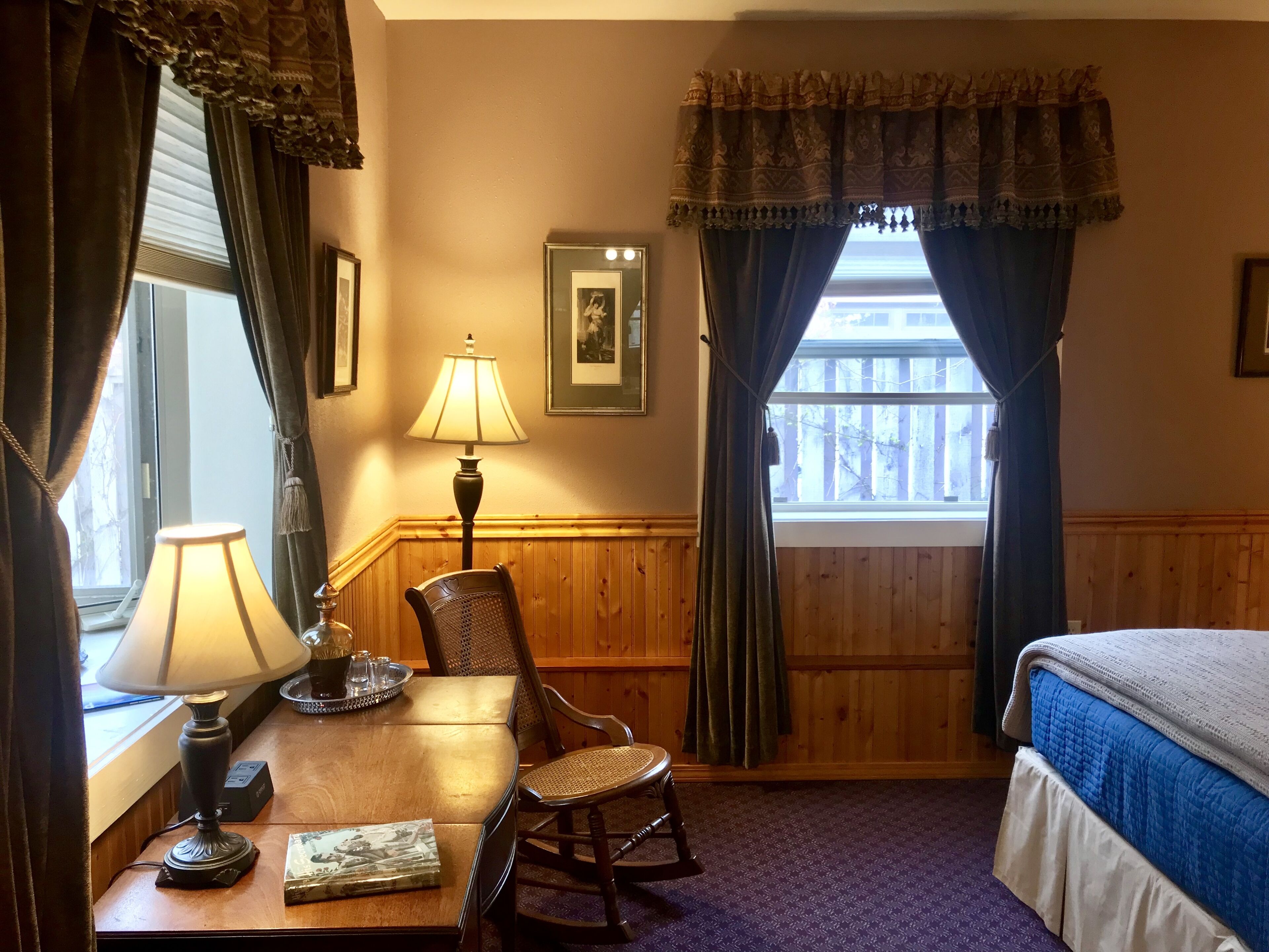 Une chambre d’hôtel avec un mur lambrissé en bois, un lit recouvert d’une courtepointe bleue, un bureau avec une lampe, une chaise et une fenêtre avec des rideaux.