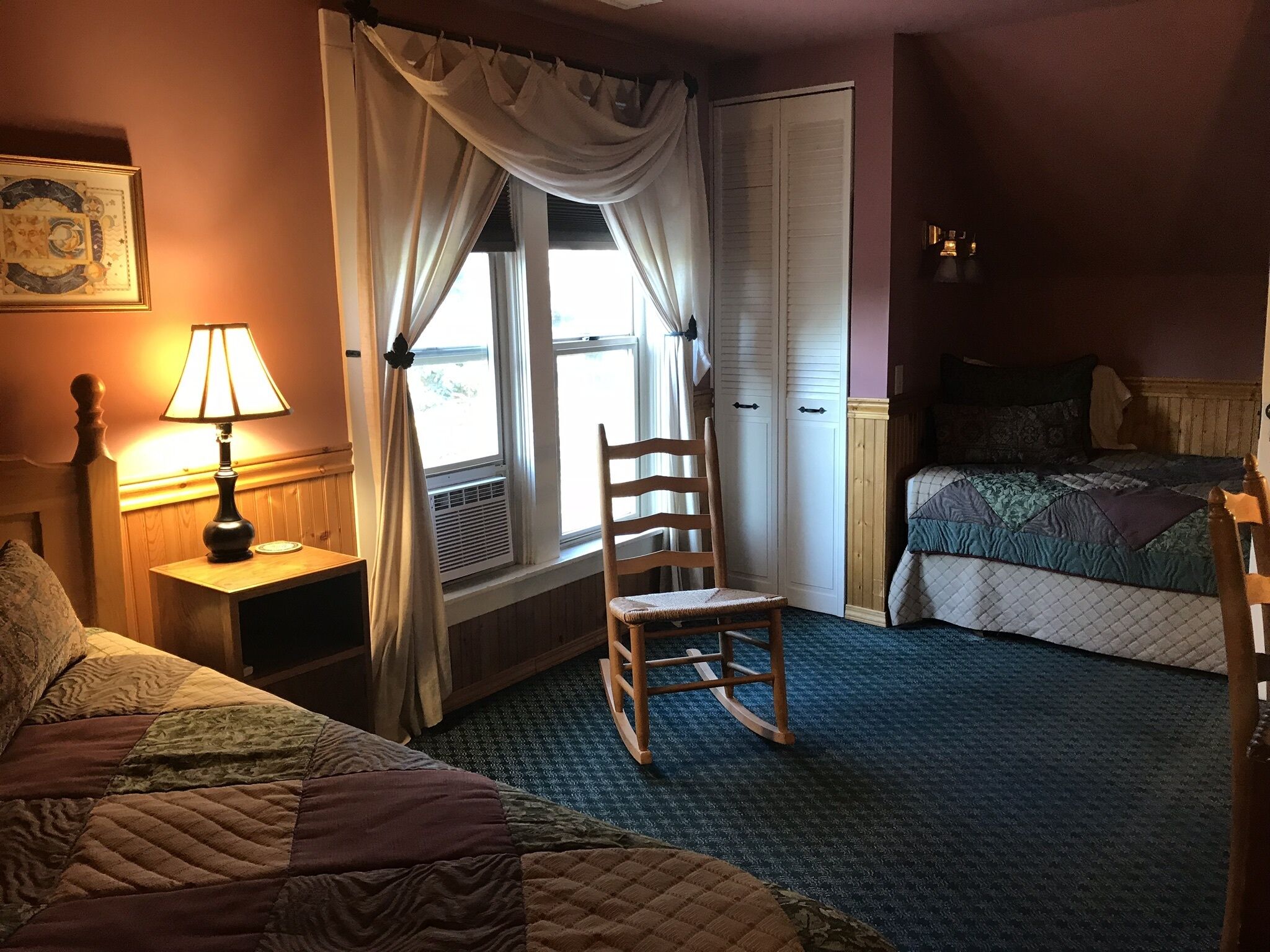 A bedroom with a bed, a rocking chair, a nightstand with a lamp, a window with curtains, and a wardrobe.