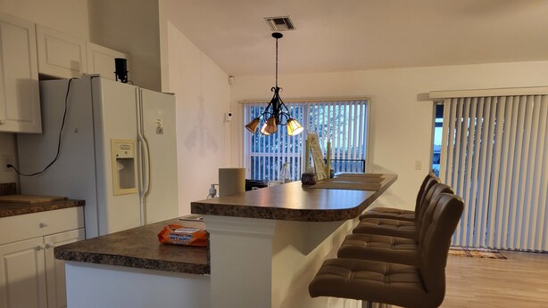 A modern kitchen with white cabinets, a granite countertop, a built-in island with bar stools, and a window with blinds. - Alabama Rd S Rancher (Lehigh Acres)