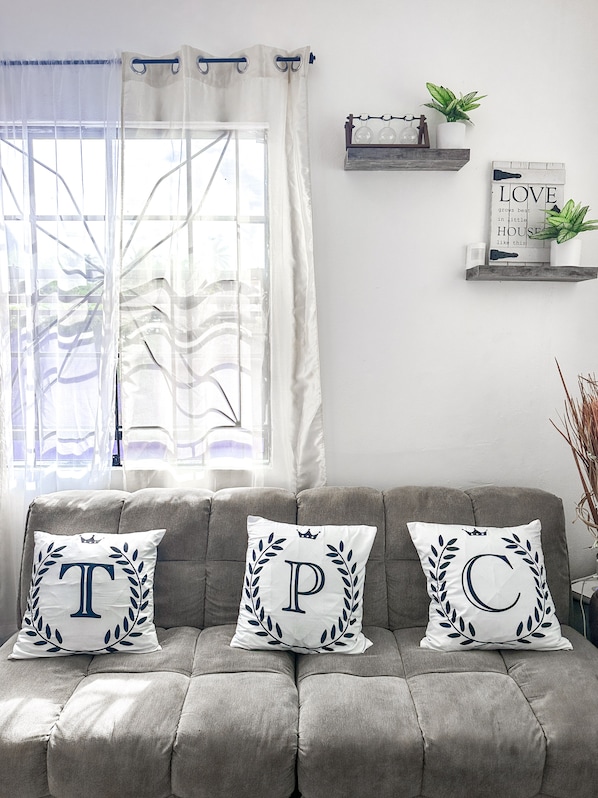 A grey sofa with three decorative pillows, a window with sheer curtains, and a wall with a shelf holding plants and a sign. - Purple Cottage, Hidden Gem near Hummingbird Beach (Soufriere)