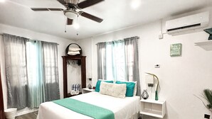 A modern bedroom with a bed, a ceiling fan, an air conditioning unit, a bedside table with a lamp, and a decorative wall piece. - Purple Cottage, Hidden Gem near Hummingbird Beach (Soufriere)