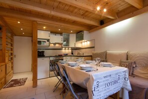 A cozy dining area with a table set for two, a kitchen in the background, and a sofa with cushions. - Grand Roc C125 280m from Slopes, View & Parking, Argentière (Chamonix), France (Argentière (Chamonix))
