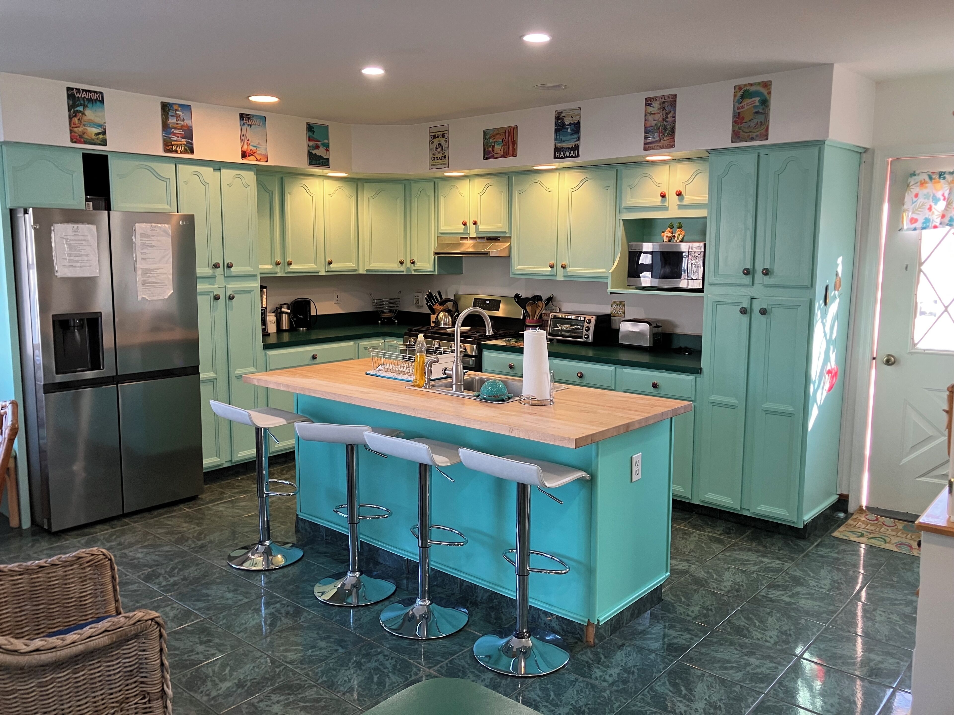 A kitchen with teal cabinets, a stainless steel refrigerator, a wooden island with bar stools, and a tiled floor.