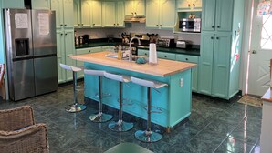 Cocina con gabinetes azul verdosos, refrigerador de acero inoxidable, una isla de madera con taburetes de bar y piso de baldosas.