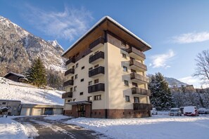 Un immeuble à plusieurs étages doté de balcons, d’un espace de stationnement et entouré d’un paysage enneigé.