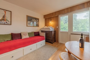 A room with a bed, a wooden dresser, a table with a bottle, and a window with a view of trees.