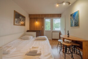 Un dormitorio con una cama, un escritorio de madera con taburetes, un radiador y dos ventanas.