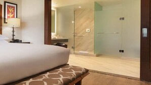 Quarto de hotel com uma cama grande, um banco, uma mesa de cabeceira com abajur, um banheiro com box de vidro e um espelho.