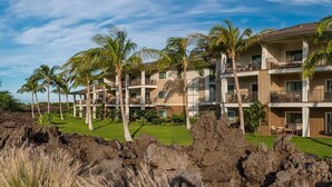 Um hotel com vários andares, varandas, palmeiras e um primeiro plano rochoso.