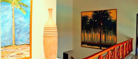 Um corredor com guarda-corpo de madeira, um quadro de uma palmeira e um vaso sobre um pedestal.