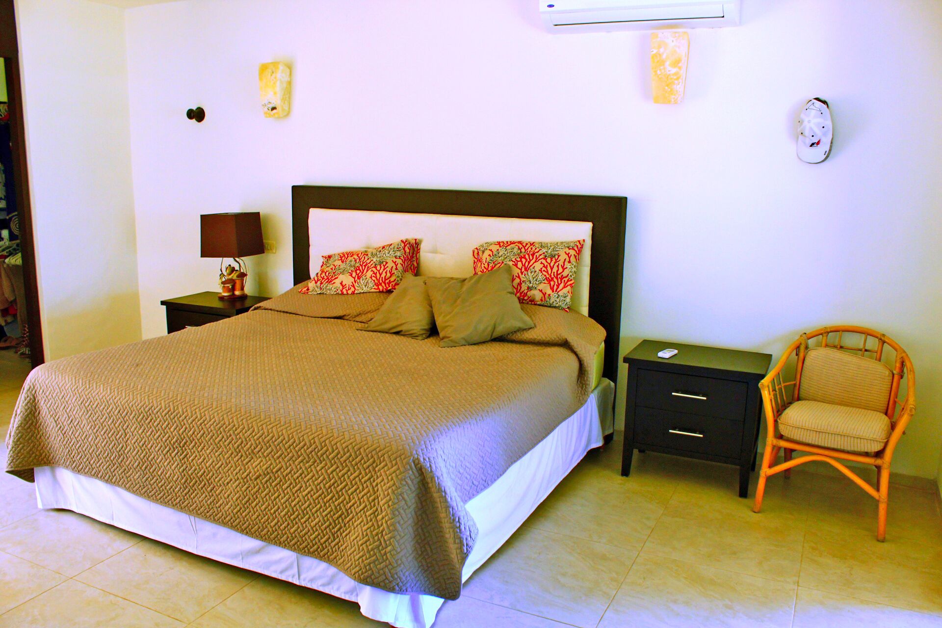 A neatly made bed with a brown bedspread, a nightstand with a lamp, a wicker chair, and a wall-mounted air conditioner.